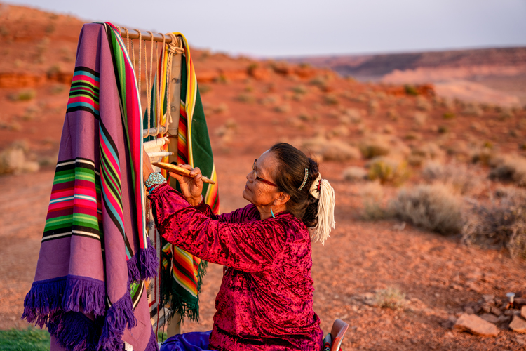 Navajo woman sitting weaving on a loom used by Navajo weavers. Fabric is multicolored in purples, reds, greens, and yellows